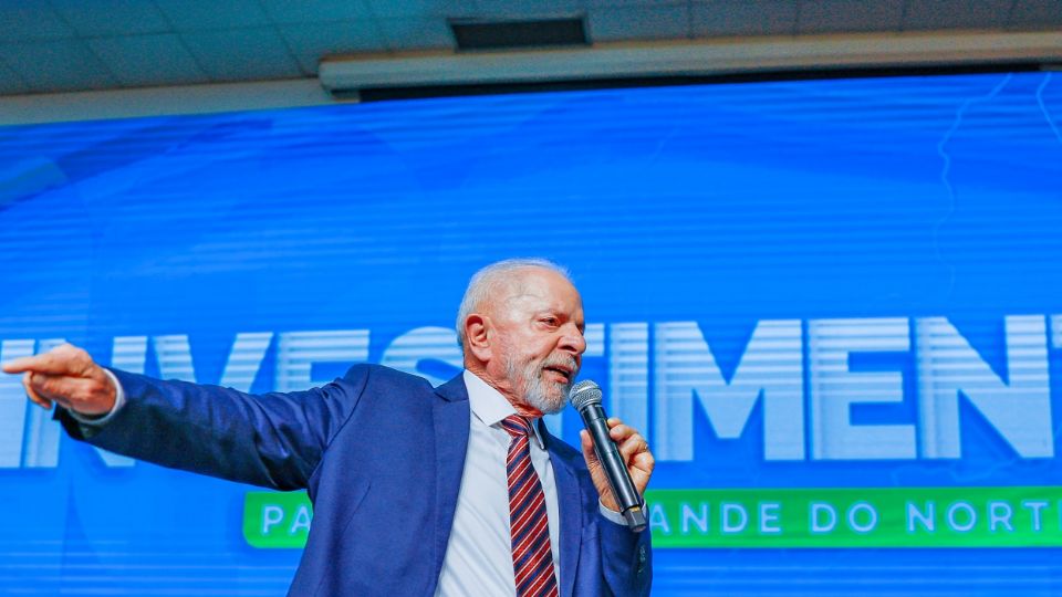 O presidente Luiz Inácio Lula da Silva discursa em um evento do governo. Ao fundo, uma tela onde se lê a palavra 'investimento'