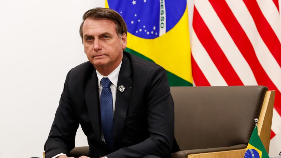 O ex-presidente Jair Bolsonaro, de terno preto, camisa branca e gravata azul escuro, sentado em uma poltrona. Ao fundo, as bandeiras do Brasil e dos Estados Unidos